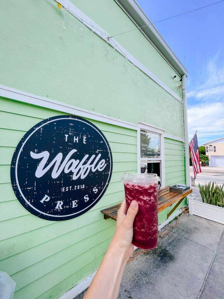 hand holding a berry smoothie in front of mint green "The Waffle Press" restaurant and sign