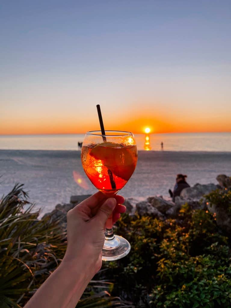 Hand holding an Aperol Spritz during sunset on the beach at The beach House on Anna Maria Island. Anna Maria Island Restaurant Guide - Where to eat on Anna Maria Island.