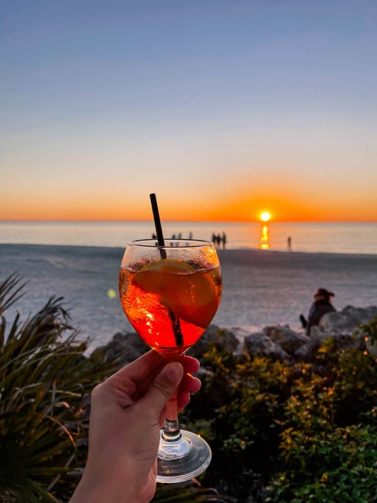 hand holding up an orange colored cocktail with the Gulf of Mexico sunset in the background